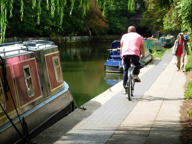 A man in a pink t-shirt is cycling on the Roachdale Canal path. To the side of him are a number of canal boats moored at the side of the canal